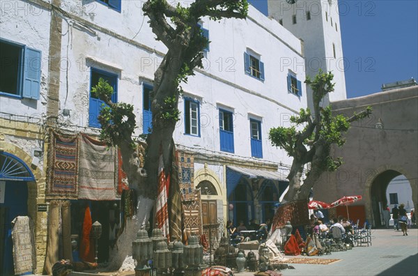 MOROCCO, Essaouira, Square with white painted buildings with blue shutters.  Shop with display of Moroccan lanterns and carpets beside cafe with striped awning and outside tables