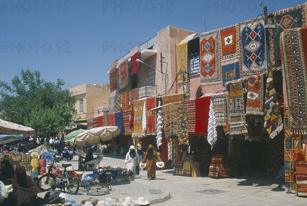 MOROCCO, Marrakesh, "The souk with carpets displayed from walls and awnings of shopfronts opposite street vendors selling shoes, hats and woven basket."