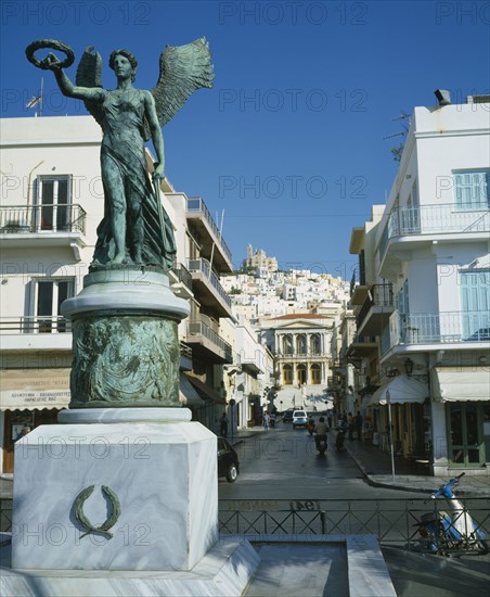 GREECE, Cyclades Islands, Syros, Ermoupolis. A statue on the seafront with the Town Hall and the Orthodox church of Anastassi on top of the hill.