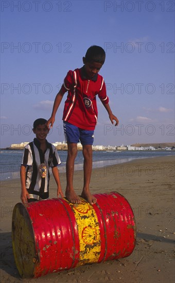 OMAN, Sur, Two young Omani boys playing on an abandoned Shell oil drum on the beach.