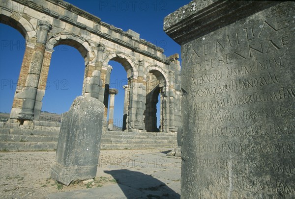 MOROCCO, Volubilis, The Basilica.  Cropped view of stone block inscribed with Roman script with arches and colonnade built into wall beyond.
