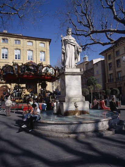 FRANCE, Aix en Provence, "Statue in centre of water fountain, people sat on edge. Carousel and buildings behind."
