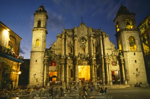 CUBA, Havana, Plaza de la Catedral.  Cathedral facade and bell tower illuminated at night with people at outside cafe tables in plaza in the foreground.