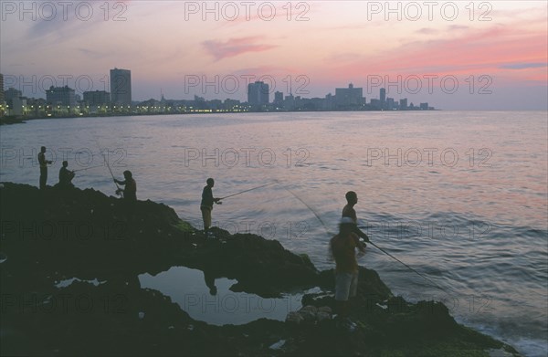 CUBA, Havana, City skyline at sunset with fishermen beside the Malecon silhouetted against pink and purple sky reflected in the sea in the foreground.