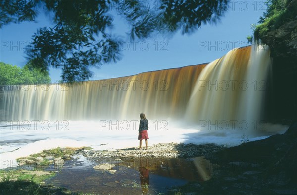 ESTONIA, Jagala, "Young woman standing in front of Jägala, steep horseshoe shaped waterfall between Tallinn and Lahemaa National Park"