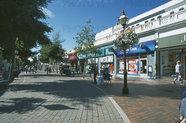 ENGLAND, East Sussex, Eastbourne, Pedestrian shopping area with line of shops and a row of small trees casting shadows along the paveing.