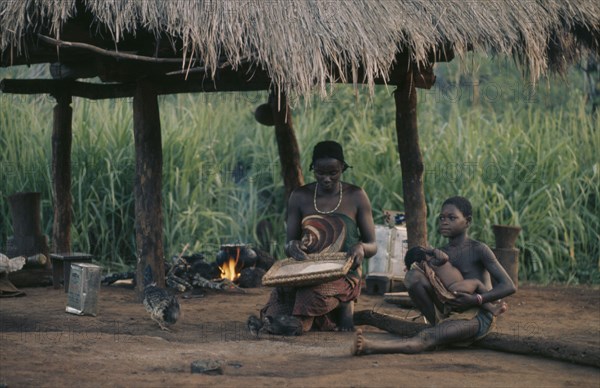 CONGO, Tribal People, Woman and children sitting beneath thatched shelter in village with pot on open fire behind.