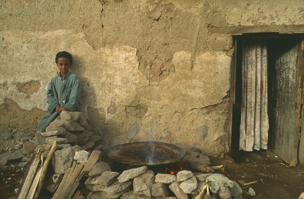 ERITREA, Asmara, Child sitting outside poor household with injera cooking on open fire at side.