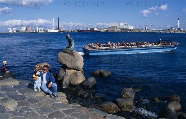 DENMARK, Zealand, Copenhagen, Family posing for photograph beside the Little Mermaid statue with passing tourist boat behind and city skyline beyond.