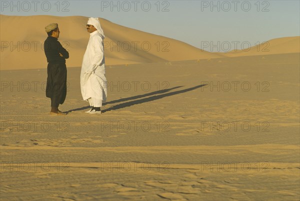 ALGERIA, Desert, Two men standing in the desert in conversation casting strong shadows across the sand.