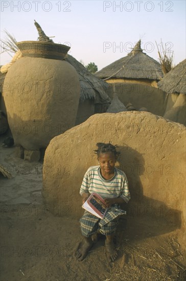 BURKINA  FASO, Bisaland, Sigue Voisin, Near Garango.Young girl called Mariam sat against wall doing her homework with grain store and village houses behind.