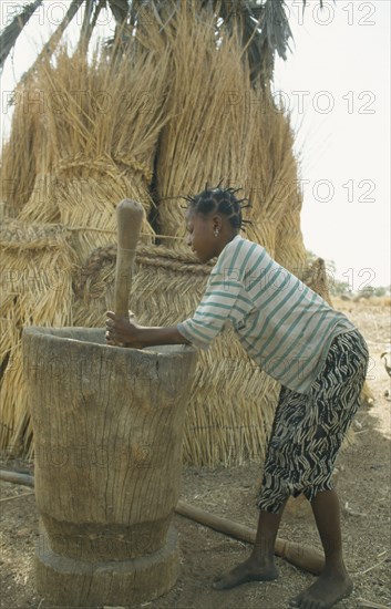 BURKINA  FASO, Bisaland, Sigue Voisin, Near Garango. Young girl pounding grain with straw roofs and granaries behind