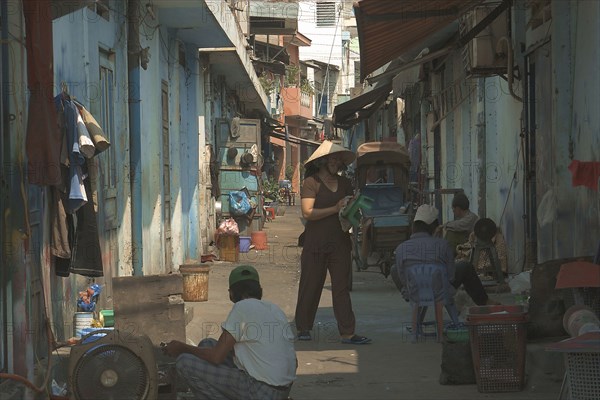 VIETNAM, South, Ho Chi Minh City, View down a side street with a woman wearing a conical hat and people sat on the ground