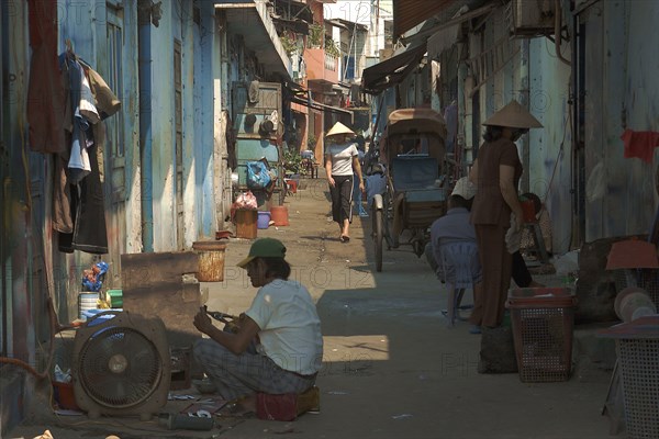 VIETNAM, South, Ho Chi Minh City, View down a side street with people sat on ground and a woman approaching wearing conical hat.