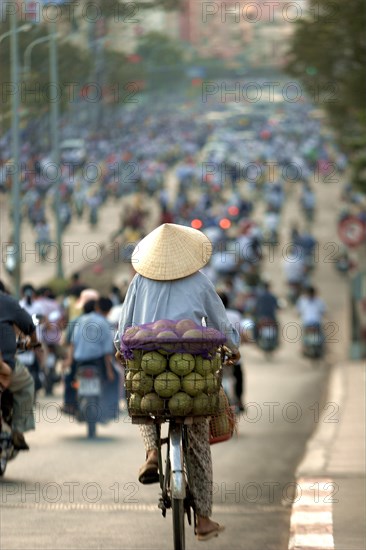 VIETNAM, South, Ho Chi Minh City, Man wearing a conical hat riding a bicycle with coconuts on the back in a busy street full of motorbikes and bicycles.
