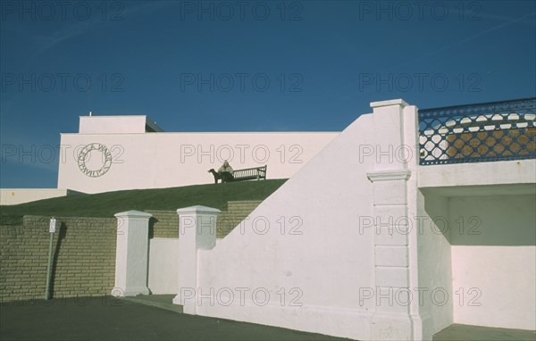 ENGLAND, East Sussex, Bexhill on Sea, De La Warr Pavilion. Exterior view from promenade towards the Pavilions white terrace and wall with the circular silver Art Deco metal sign with a woman sat on bench.