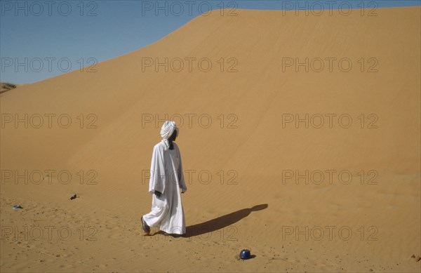 SUDAN, Kordofan Province, El Humra Village, Man in white robe and turban passing huge sand dunes encroaching on village and agricultural land and cutting off road to El Caeid.