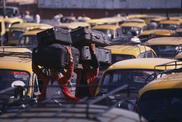 20073434 INDIA  Delhi Porters carrying luggage on their heads through parked taxi cabs at New Delhi railway station.