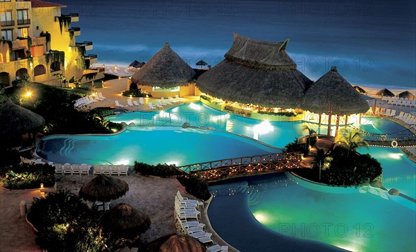 MEXICO, Jalisco ,  Puerto Vallarta, "A swimming pool surrounded by sun loungers, wooden thatched huts and bridges lit up at night. Hotel right next to beach."