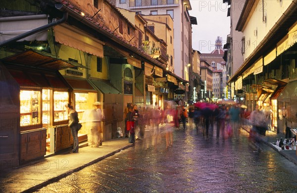 ITALY, Tuscany, Florence, "Street scene at dusk with shop fronts lining Ponte Vecchio, crowds in blurred movement and lights reflected on wet paving."