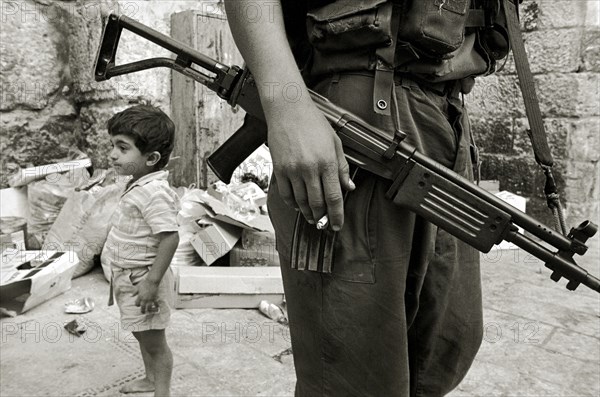 ISRAEL, Jerusalem, "Israeli soldier with rifle and cigarette relaxes in the Arab Quarter, small boy standing behind."
