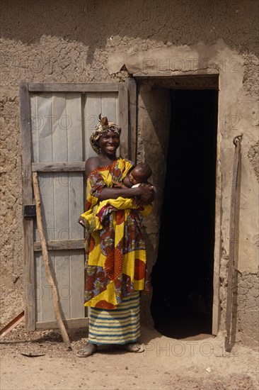 MALI, People, Portrait of Malian mother and breastfeeding baby standing outside open doorway of mud brick building.