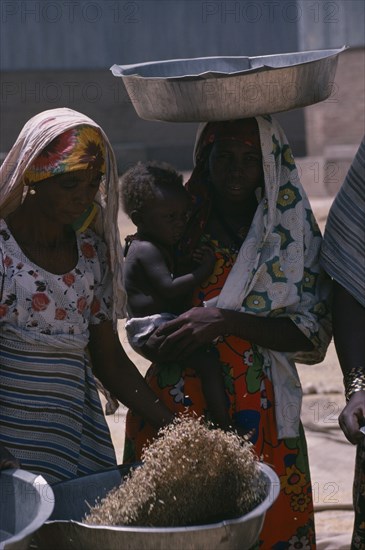 SUDAN, Work, Nigerian woman winnowing grain beside woman carrying child in her arms and large dish on her head.