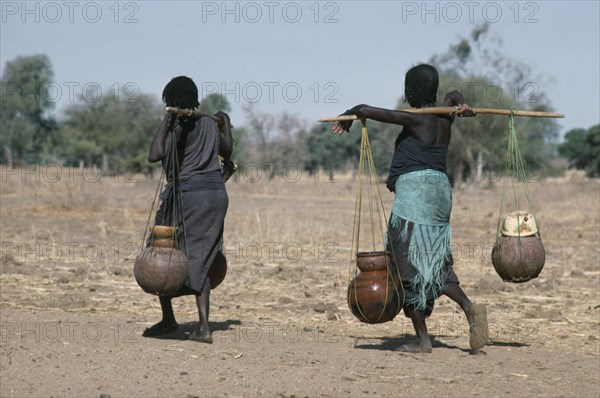 SUDAN, Habila Settlement, Chadian refugee women carrying water vessels hanging from pole across their shoulders from well in settlement near El Geneina.