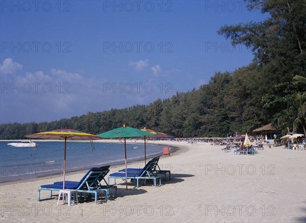 THAILAND, North Phuket, Naiyang Beach, "View along the sandy beach with sun loungers and colourful parasols in the foreground and a row of them all along the water’s edge.. Red sign for boat hire and people swimming, boat in the water."