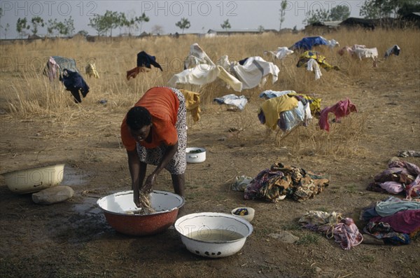 GHANA, Laundry, Laundress washing clothes in bowls with garments spread out over bushes to dry behind.