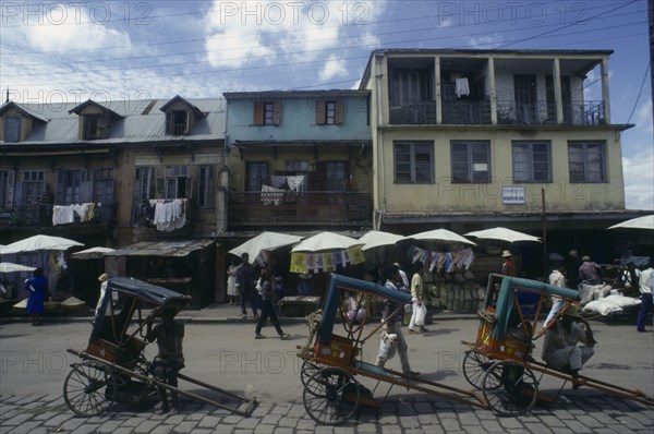 MADAGASCAR, Antananarivo, "Street scene with washing hanging from buildings above roadside stalls and pousse pousse drivers waiting for customers.  People walking past, some with bags."