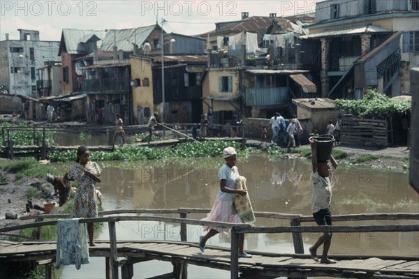 MADAGASCAR, Antananarivo, Children crossing wooden bridge over stagnant water in poor housing area near Antananarivo.