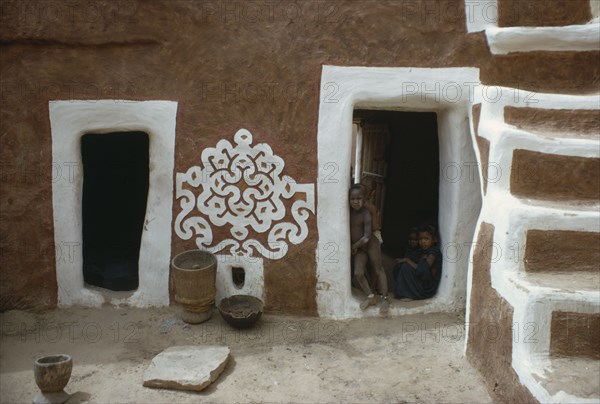 MAURITANIA, Oualata, Traditional mud architecture decorated with bas relief motif of applied gypsum  white and red clay.  Children framed in open doorway with white surround.