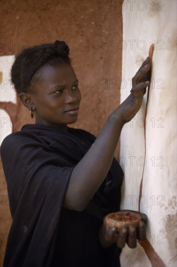 MAURITANIA, Oualata, Young woman decorating exterior wall of mud building with ochre.  It is traditional for women to both design and apply such decorations.