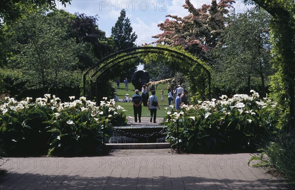 ENGLAND, Surrey, Woking, Wisley Royal Horticultural Society Garden. Visitors walking along pathway through arch towards Henry Moore sculpture.