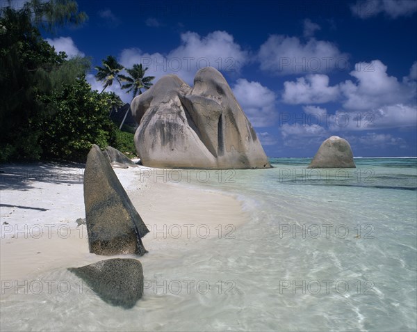 SEYCHELLES, La Digue, Source De Argent, Large rock formations on sandy beach with clear shallow water and palm trees growing along coastline