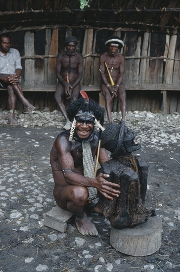 INDONESIA, Irian Jaya, Baliem Valley, Dani Warrior man holding smoked ancestral mummy with three men sat behind him looking on