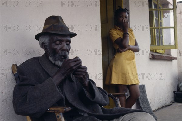 SOUTH AFRICA, Gauteng, Alexandra Township, Portrait of elderly seated man lighting pipe with girl standing in doorway behind.