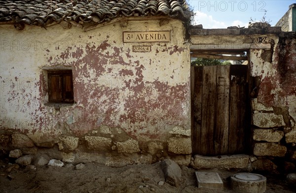 GUATEMALA, Quetzatenango, Xela, "A back street in the outskirts of the Guatemalan town of Xela. A characteristic but crumbling house sits baking in the sun surrounded by large rocks and a dry dusty road. The house itself is worn and crumbling. Some of the tiles on the roof are beginning to crack and the old red brickwork can be seen behind flaking paint. On the front wall of the house hangs the road and area name - '3 Avenida, Zona 4' The entrance to the house consists of an old but firm wooden gate. The gate, made of odd lengths of wood is closed but one can just make out the green foliage in the background. The sky is mainly clear and blue and to the left of the road name sits a small hole in the front of the house with a closed shutter in."