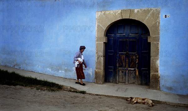 GUATEMALA, Quetzatenango, Xela, "A back street in the outskirts of the Guatemalan town of Xela. A characteristic but slightly crumbling house sits baking in the sun in front sits a dry dusty road. The house itself is a little worn but glows a beautiful blue colour in the afternoon sun. On the front wall of the house sits the main entrance, a fairly grand double wooden door underneath proud stone archway. To the right sits the number of the house. An old indigenous woman slowly walks past the door of this house, dressed as a country person or 'campesino'. She carries a bundle of clothes and wears a cotton headdress to protect her from the strong sun. On the dusty ground infront of the house lies a lazy stray dog, taking advantage of the shade and the cool ground."