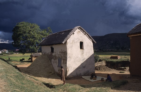MADAGASCAR, Architecture, Road to Ambositra. Women outside thatched white building with straw in a pile next to the wall of the building and dramatic dark cloud formation in the sky