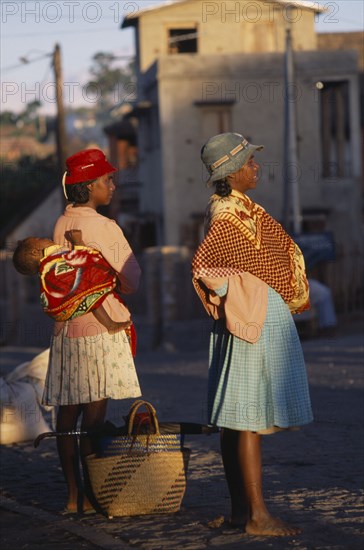 MADAGASCAR, People, Women, Road to Fianarantsoa. Two women one carrying a baby in a sling on her back wearing colourful clothing standing waiting for a lift with golden sunlight shining onto them
