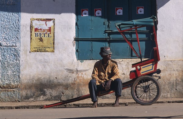 MADAGASCAR, Ambalavao, Man sitting on his rickshaw awaiting a fare at the side of the road with a building behind