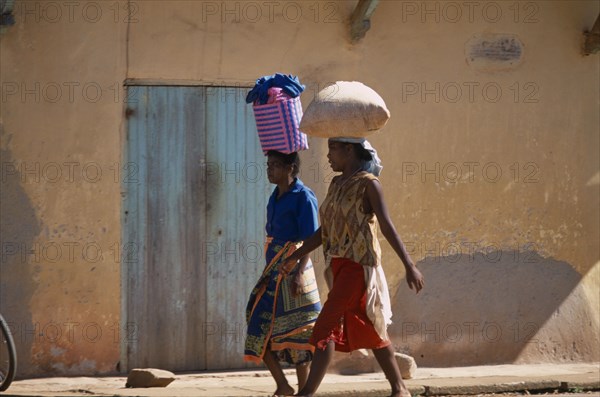 MADAGASCAR, Ambalavao, Two women wearing colourful clothing walking along a street next to a building one carrying a shopping basket on her head and the other woman carrying a bag of grain