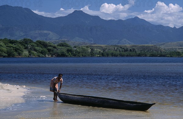 MADAGASCAR, Fort Dauphin, Lokaro, A woman standing at the waters edge of a sandy beach about to push her Pirogue back into the water to return to her village with mountains in the distance