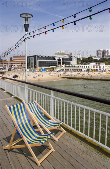 ENGLAND, Dorset, Bournemouth, Deckchairs on the Pier withThe East Beach and the Imax Complex on the left beside seafront restaurants and bars. Adults and children play in the sand on the beach and at the water's edge between the groynes. Hotels and flats line the clifftop into the distance