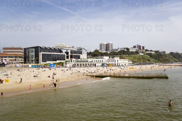 ENGLAND, Dorset, Bournemouth, The East Beach with seafront restaurants and bars. Adults and children play in the sand on the beach and at the water's edge between the groynes. Hotels and flats line the clifftop into the distance