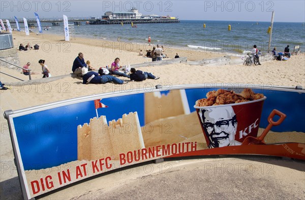 ENGLAND, Dorset, Bournemouth, Tourists playing in the sand and watching speedboat races off the West Beach beside the pier. A KFC advertising board in the foreground.