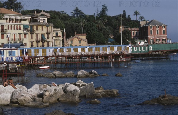 ITALY, Liguria, Rapallo, Coastal town on the Italian Riviera with line of changing huts on jetty out into rocky bay wand waterside buildings behind.