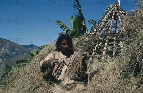 COLOMBIA, Sierra Nevada de Santa Marta, Northern Slopes, Mama Valencia a Kogi mama/priest re-thatching the conical roof of his house with recently cut hay in lowland settlement of San Antonio He wears traditional criss-crossed mochilas/ shoulder bags over a woven cotton manta/cloak
